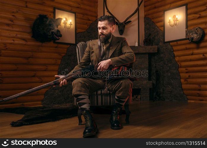 Hunter man in vintage hunting clothing sitting in a chair with antique rifle. Fireplace, stuffed wild animals, bear skin and other trophies on background. Hunt lifestyle