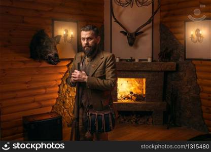 Hunter man with old gun in vintage traditional hunting clothing standing against antique chest. Fireplace, stuffed wild animals, bear skin and other trophies on background. Hunter man with old gun against antique chest