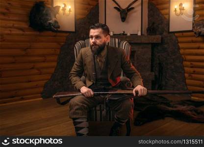 Hunter man with old gun in vintage traditional hunting clothing standing against antique chest. Fireplace, stuffed wild animals, bear skin and other trophies on background. Hunter man with old gun against antique chest