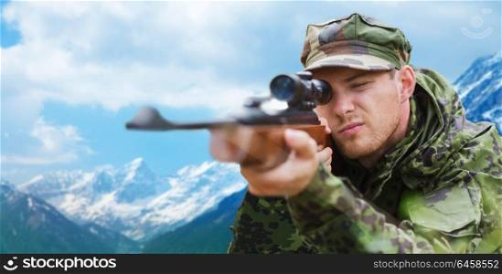 hunting, army, military service and people concept - young soldier, sniper or hunter with gun aiming or shooting over mountains background. soldier or hunter with gun aiming or shooting