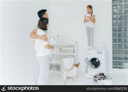 Husband and wife embrace, stand in laundry room, look happily at their single child who stands on washing machine with bottle of washing powder, wash clothes, do family domestic chores at weekend