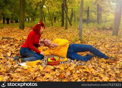 husband and wife in bright clothes for relaxing settled down on a picnic among the trees and fallen leaves with a bottle of wine and glasses