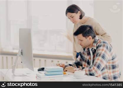 Husband and wife work on paper report together, check information, sit in front of computer, drinks coffee, dressed in casual wear, collaborate for teamworking. Family and remote work concept
