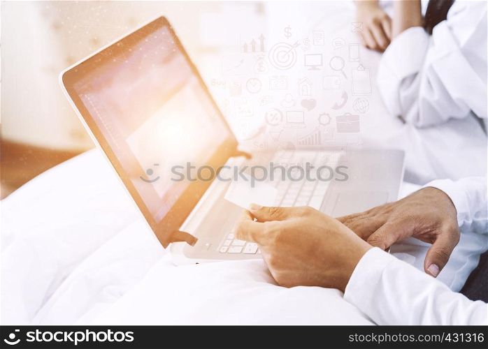 Husband hand holding a credit card and using a laptop for working or online purchasing on bedroom in the morning at home. Online business concept.