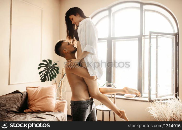 Husband holding wife in his arms against window with sunlight, erotica in bedroom. Erotic scene, sexual relationship of love couple