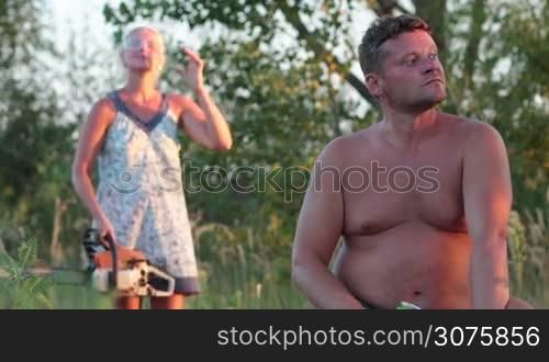 Husband sitting on a dry wood and drinking beer, behind his back smiling wife with working chainsaw standing and joking with him during summer vacation
