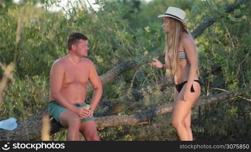 Husband sitting on fallen dry tree and drinking a beer during summer vacation. Wife in swimsiut scolds her husband for abusing alcohol and slaps his face