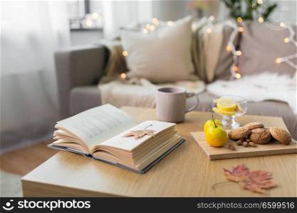 hygge and cozy home concept - book, autumn leaves, cup of tea with lemon, almond nuts and oatmeal cookies on table. book, lemon, tea and cookies on table at home