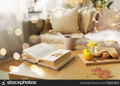 hygge and cozy home concept - book, autumn leaves, cup of tea with lemon, almond nuts and oatmeal cookies on table. book, lemon, tea and cookies on table at home