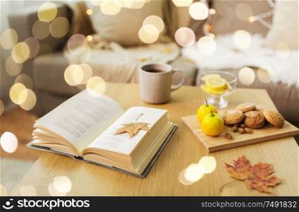 hygge and cozy home concept - book, autumn leaves, cup of tea with lemon, almond nuts and oatmeal cookies on table. book, lemon, tea and cookies on table at home