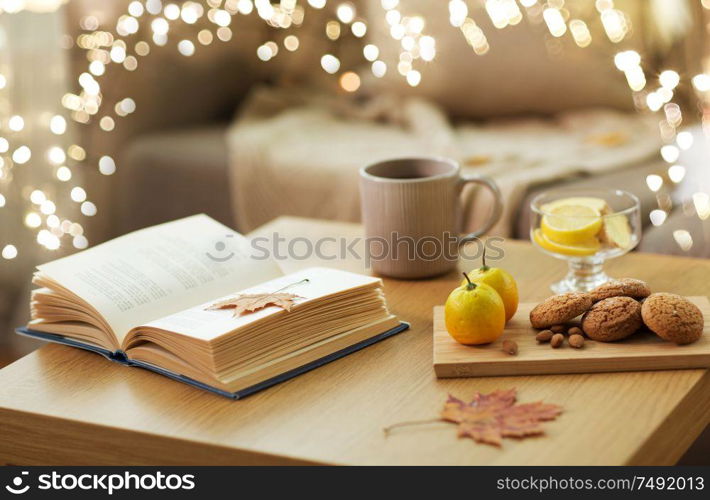 hygge and cozy home concept - book, autumn leaves, cup of tea with lemon, almond nuts and oatmeal cookies on table. book, lemon, tea and cookies on table at home