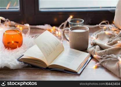 hygge and cozy home concept - book, cup of coffee or hot chocolate and candles with garland on window sill. book and coffee or hot chocolate on window sill