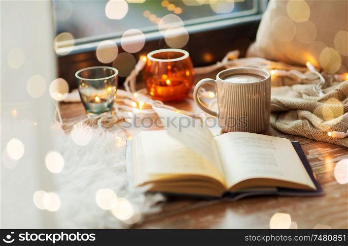 hygge and cozy home concept - book, cup of coffee or hot cchocolate and candles with garland on window sill. book and coffee or hot cchocolate on window sill