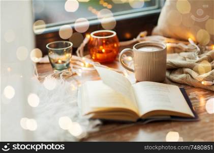 hygge and cozy home concept - book, cup of coffee or hot cchocolate and candles with garland on window sill. book and coffee or hot cchocolate on window sill
