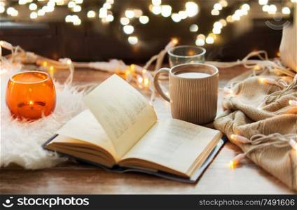 hygge and cozy home concept - book, cup of coffee or hot chocolate and candles with garland on window sill. book and coffee or hot chocolate on window sill