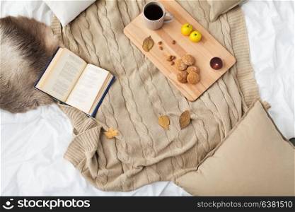 hygge and cozy home concept - cookies, lemon tea, book and leaves in bed. cookies, lemon tea, book and leaves in bed
