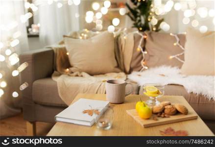 hygge and cozy home concept - cup of tea with lemon, book, autumn leaves and oatmeal cookies on wooden table in living room. tea with lemon, book and cookies on table at home