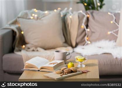 hygge and cozy home concept - oatmeal cookies, book, tea and lemon on wooden table in living room. oat cookies, book, tea and lemon on table at home