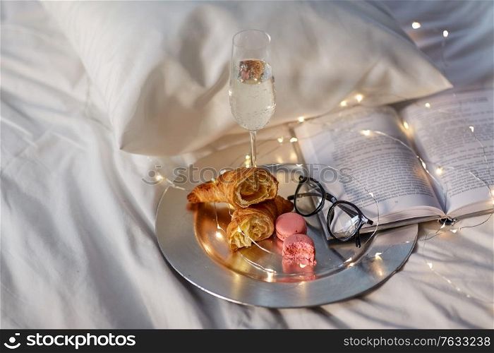 hygge and lifestyle concept - glass of champagne wine, croissants, macaroons with book and glasses in bed at home. champagne, croissants, book and glasses in bed
