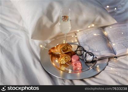 hygge and lifestyle concept - glass of champagne wine, croissants, macaroons with book and glasses in bed at home. champagne, croissants, book and glasses in bed