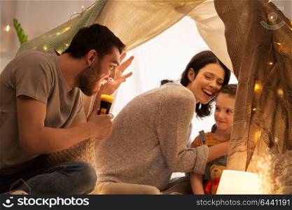 hygge and people concept - father with torch light telling scary stories to his daughter and wife, family having fun in kids tent at night at home. father telling scary stories to his daughter