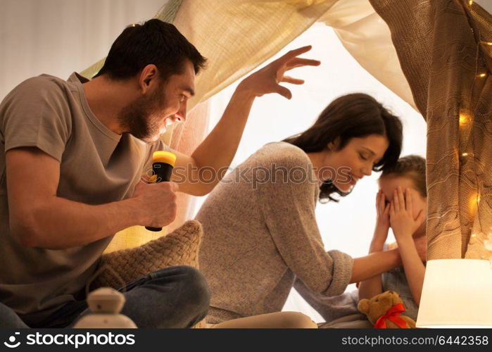 hygge and people concept - father with torch light telling scary stories to his daughter and wife, family having fun in kids tent at night at home. father telling scary stories to his daughter