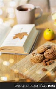 hygge, bake and food concept - oatmeal cookies, almonds, book and tea on wooden table at home. oat cookies, almonds and book on table at home
