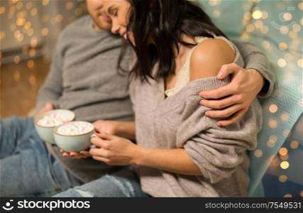 hygge, christmas and holiday concept - close up of happy couple drinking hot chocolate with marshmallow at home over shimmering festive lights. close up of couple with hot chocolate at home