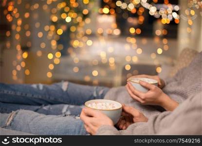 hygge, leisure and christmas concept - close up of couple drinking hot chocolate with marshmallow at home over shimmering festive lights. close up of couple drinking hot chocolate at home