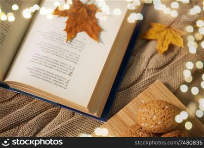 hygge, literature, reading and poetry concept - book with autumn leaf, lemons and oatmeal cookies on sofa. book with autumn leaf and cookies on home blanket