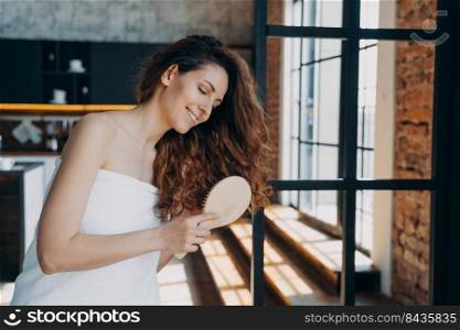Hygiene and hair care concept. Young european woman combs her long gorgeous curly hair with brush. Happy girl wrapped in towel doing hair therapy in modern room of apartment or hotel.. Hygiene and hair care concept. Young european woman combs her long gorgeous curly hair with brush.