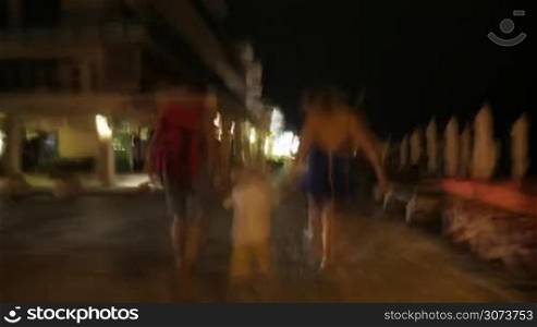 Hyperlapse shot of family walking along the night street in resort town, they passing by stores, cafes and hotels. Then child going on sidewalk alone