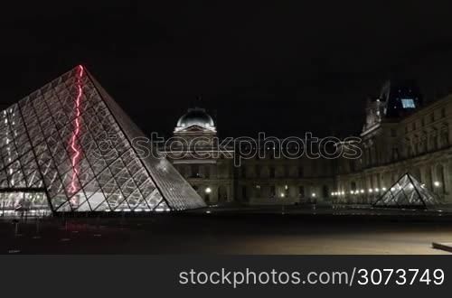 Hyperlapse shot of few people walking outside the Louvre and illuminated glass Pyramid at night. World famous art museum in Paris, France