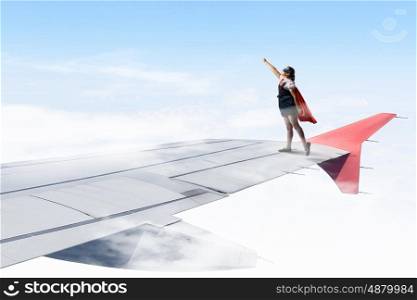 I am a super girl. Powerful super hero kid girl standing on wing of flying airplane