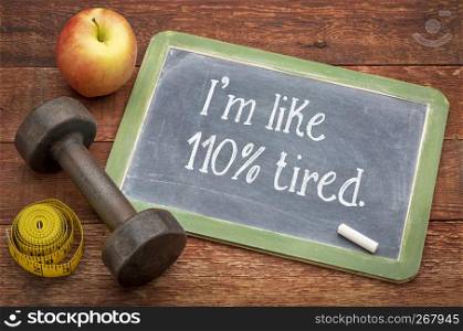 I am like 110% tired - white chalk text on a slate blackboard against weathered red painted barn wood with a dumbbell, apple and tape measure