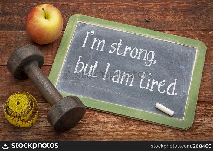 I am strong, but I am tired - white chalk text on a slate blackboard against weathered red painted barn wood with a dumbbell, apple and tape measure