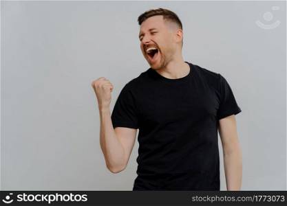 I am winner. Young excited and very emotional bearded man in black t shirt celebrating victory, clenching fist and screaming while standing against grey background. Success concept. Young overjoyed man celebrating success while standing isolated over grey background