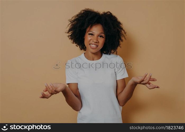 I dont know. Studio shot of confused young african female can not make decision, shrugging shoulders and does not know what happened, dressed in white t shirt feeling embarrassed. Doubtfulness concept. Confused african female can not make decision, shrugging shoulders isolated on beige background