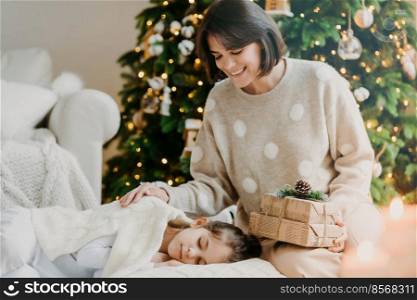 I have little secret for you. Lovely mother and daughter celebrate New Year at home. Mom poses near sleeping girl with presents against decorated New Year tree. Happy winter holidays concept
