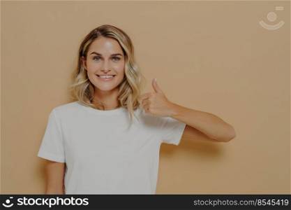 I like that. Happy young blonde female wearing white casual tshirt making thumbs up gesture and smiling cheerfully at camera, posing against yellow background. Body language concept. Happy young blonde female wearing white casual tshirt making thumbs up gesture and smiling cheerfully