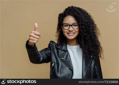 I totally agree with you. Pleased curly haired woman shows thumb up, rates excellent product, recommends promo offer, wears glasses and leather jacket, isolated on beige background, approves best deal