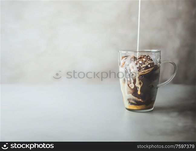 Ice coffee preparation. Cream milk pouring in glass with coffee ice cubes in glass mug on rustic table at concrete wall background. Iced coffee making. Summer refreshing beverage. Cold drink.