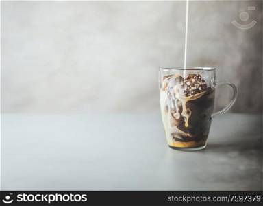 Ice coffee preparation. Cream milk pouring in glass with coffee ice cubes in glass mug on rustic table at concrete wall background. Iced coffee making. Summer refreshing beverage. Cold drink.