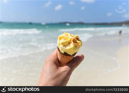 Ice cream cone in hand with sea background / Melting ice cream on beach in summer hot weather ocean landscape nature outdoor vacation , Yellow ice cream mango with nuts