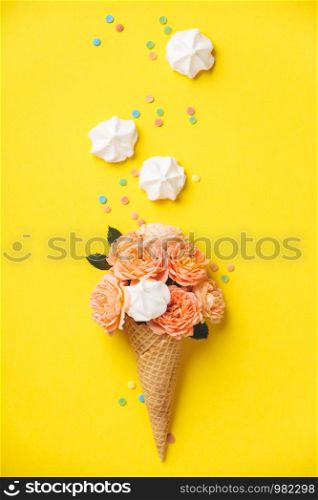 Ice cream cone with pink roses and merengues on yellow background. Summer minimal concept. Flat lay.