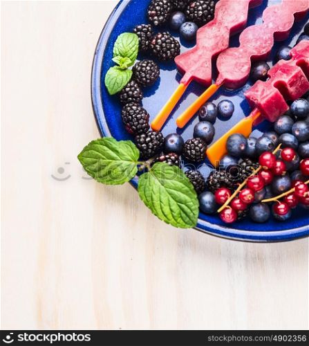Ice cream sorbet pops with different summer berries and mint in blue plate on white wooden background