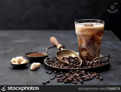 Iced black coffee with milk on tray with coffee beans and ground coffee with cane sugar and vintage soop on black background. Space for text