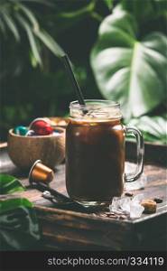 Iced coffee and coffee capsules on tropical leaves background. Iced coffee and tropical background, close up