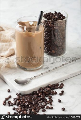 Iced cold caramel coffee with milk and glass container of beans and spoon on marbel board and light table background.