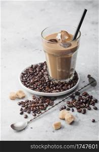 Iced cold coffee with milk and raw beans and spoon with coffee beans in white plate on light table background with cane sugar.